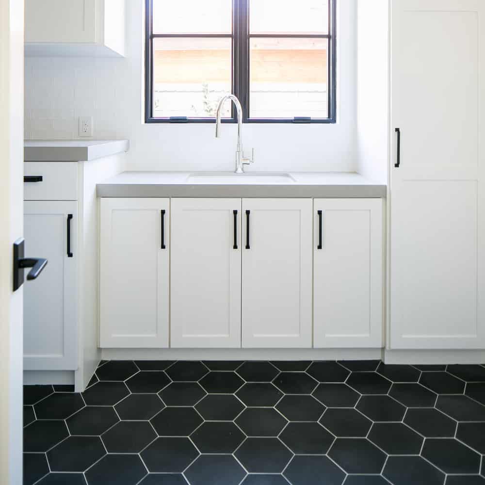 A modern kitchen features white cabinets with black handles, a light gray countertop, a stainless steel faucet, and a black Zia Tile Lattice 3 floor. Two windows above the sink provide natural light.
