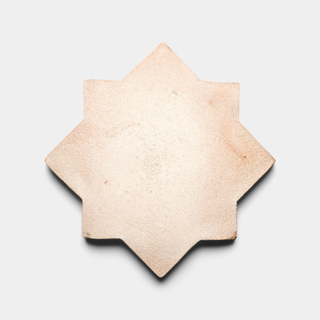 The Stars & Cross + Blanco tile by Zia Tile is a handmade beige terra cotta piece with eight pointed tips and a smooth surface, shown on a plain white background. Shadows accentuate its three-dimensional, star-shaped design.