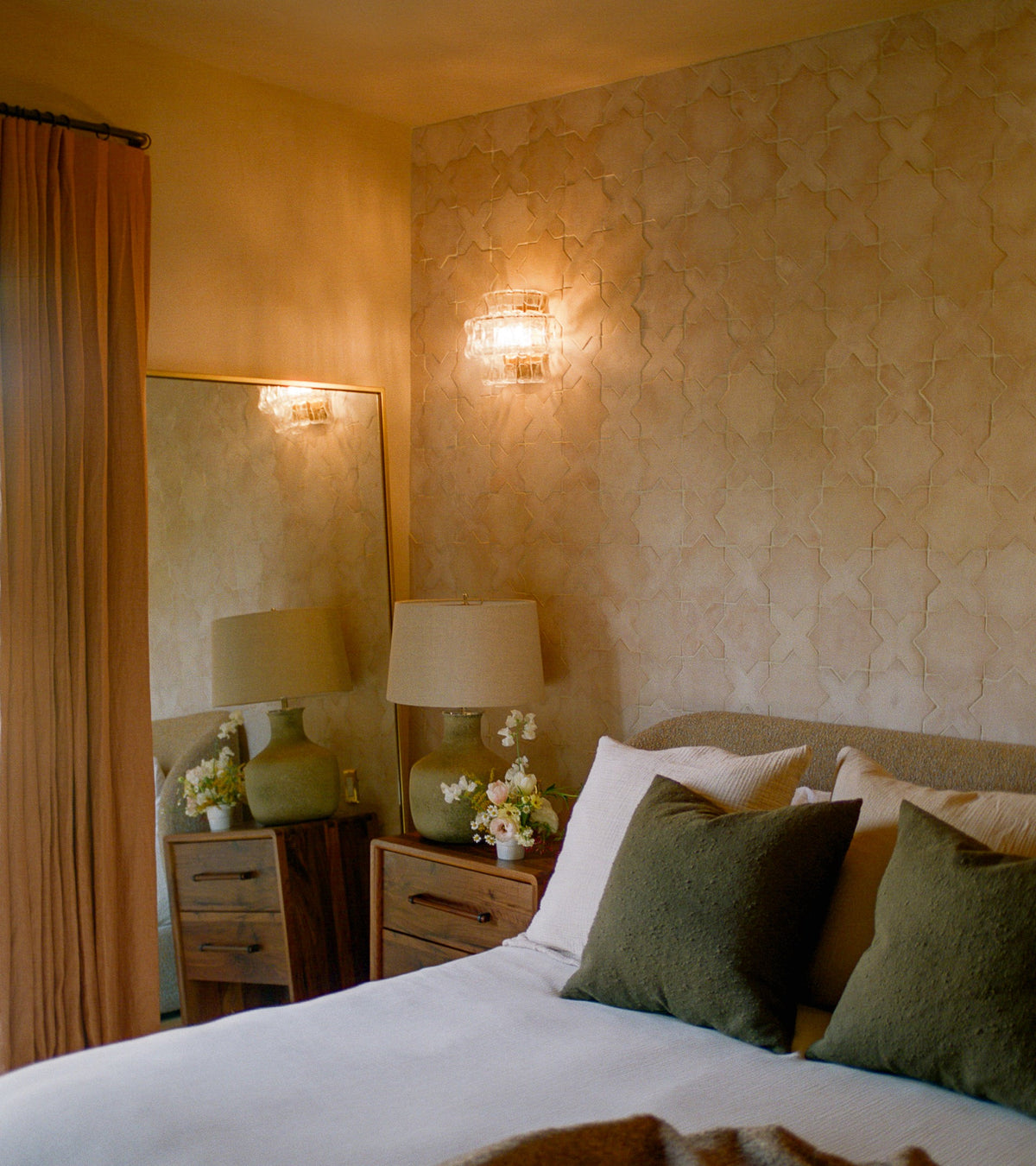 A cozy bedroom featuring Zia Tile’s Stars & Cross + Blanco tiles, white linens with green pillows, wooden nightstands, a large mirror, peach curtains, and a lit sconce reflects the handmade charm of San Miguel de Allende.