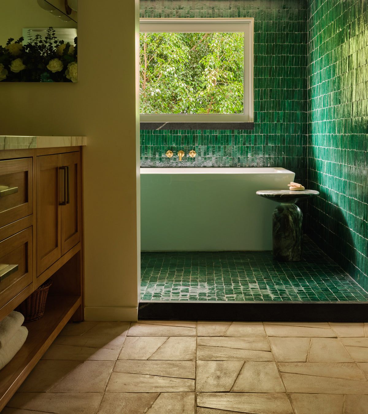 A bathroom featuring Toltec + Blanco tiles by Zia Tile in the shower and bathtub area, handmade Cotto accents, a small round side table, large window with tree views, and a wooden vanity on beige tiled floors.