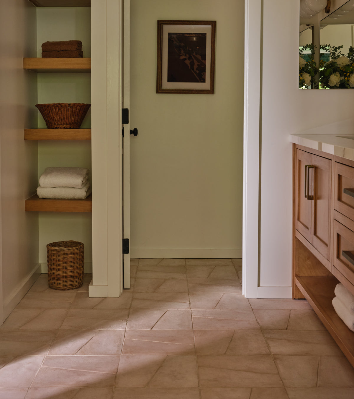 A bathroom features Zia Tile’s Toltec + Blanco handmade tiles on the floor, wooden shelves with towels and baskets, a right-side vanity with drawers, and a framed picture above the door.