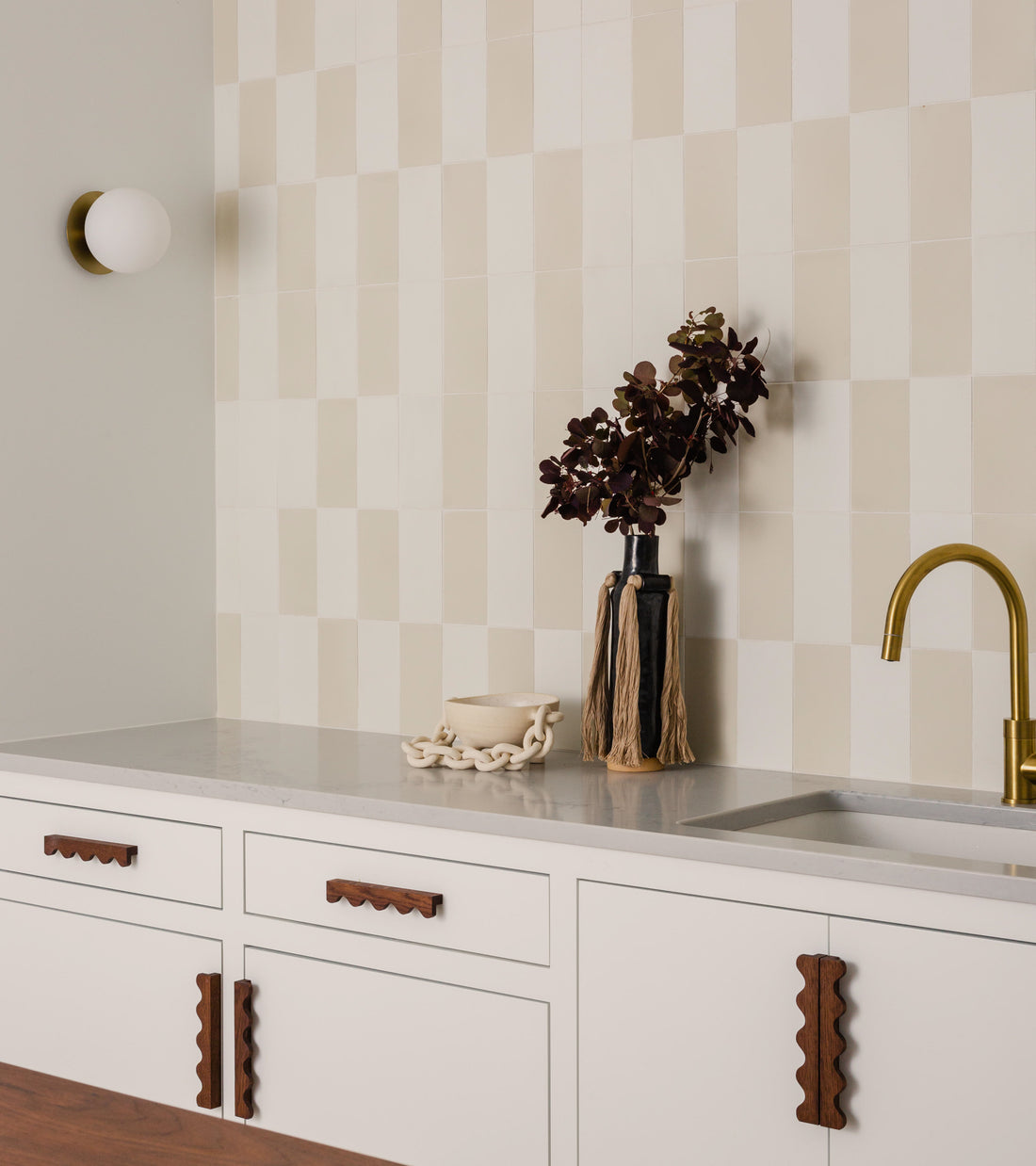 Minimalist kitchen with white cabinets, wavy wood handles, gold faucet, and light gray countertop. The backsplash features vendor-unknown’s White 4x8 tile in a checkerboard pattern. A vase with dark flowers sits by the sink.
