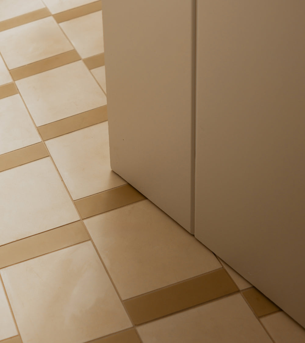 A close-up of vendor-unknown’s Bone 8x8 cement tiles in a matte finish, featuring a subtle grid pattern and alternating light beige tones, installed on the floor beside a smooth, off-white wall or cabinet.