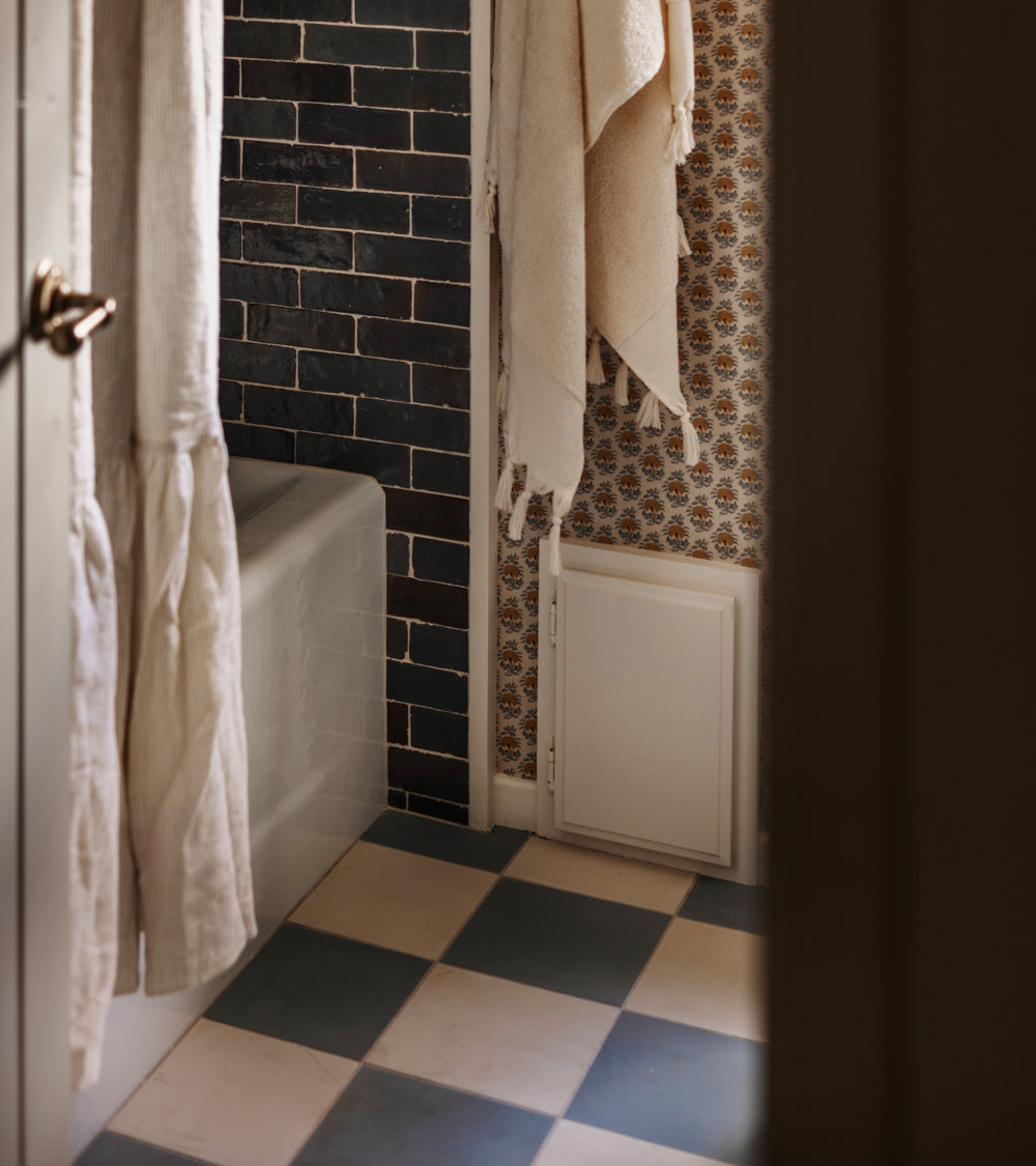 A bathroom with checkered blue and white floor tiles, Zia Tile's Graphite Grey 2x6 tiles by the tub, patterned wallpaper, and light-colored towels hanging on hooks.