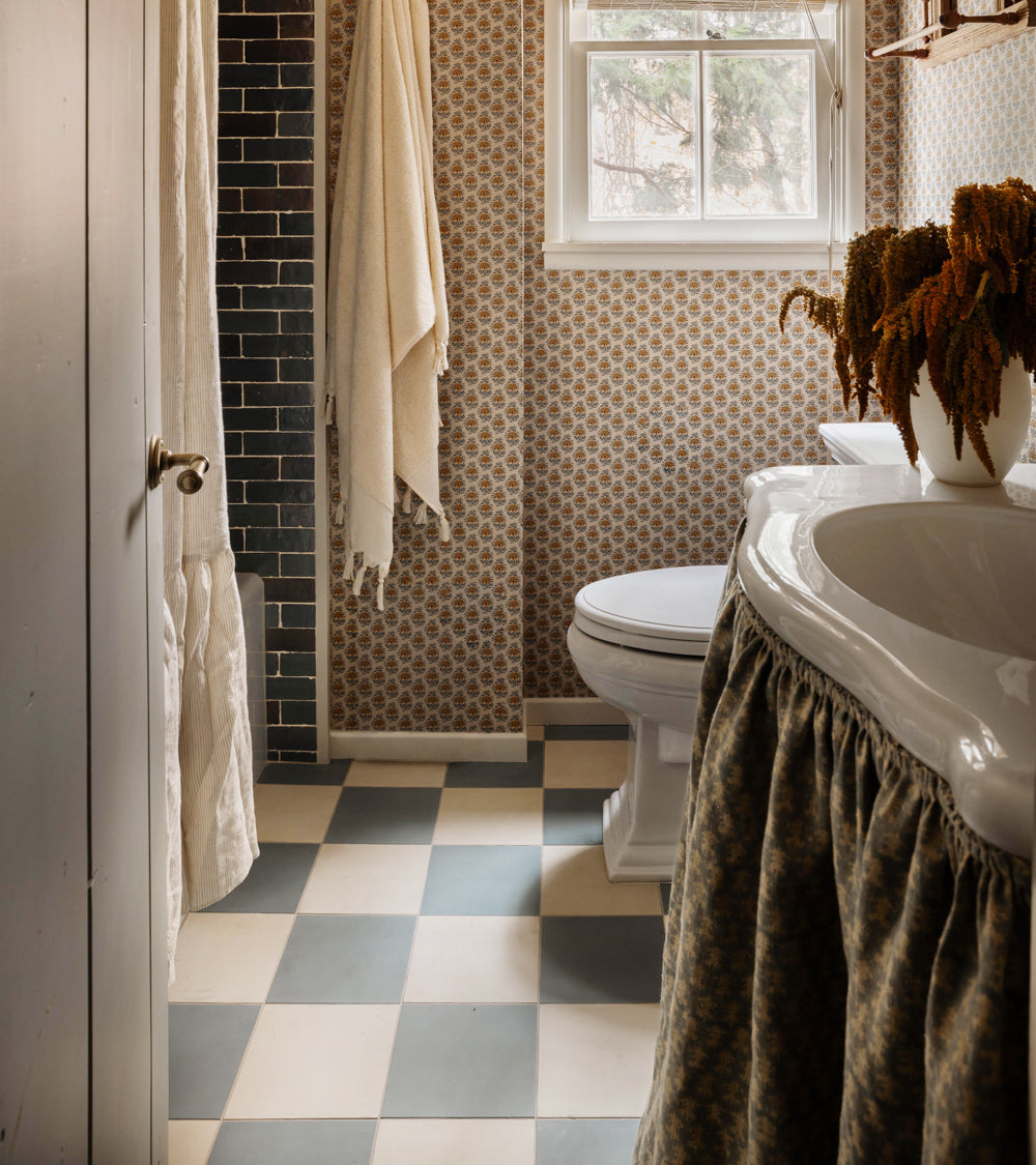A cozy bathroom features vendor-unknown Zeppelin 8x8 checkered green cement tile floor, patterned wallpaper, a white toilet, window, towel, and a white sink with floral skirt and dried flowers.