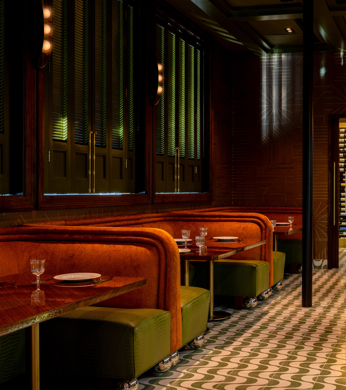 A stylish restaurant interior features plush orange booths, green cushions, marble tables with place settings, Zia Tile’s Bossa Nova Saguaro 12x12 geometric cement tile floors, and soft ambient lighting.
