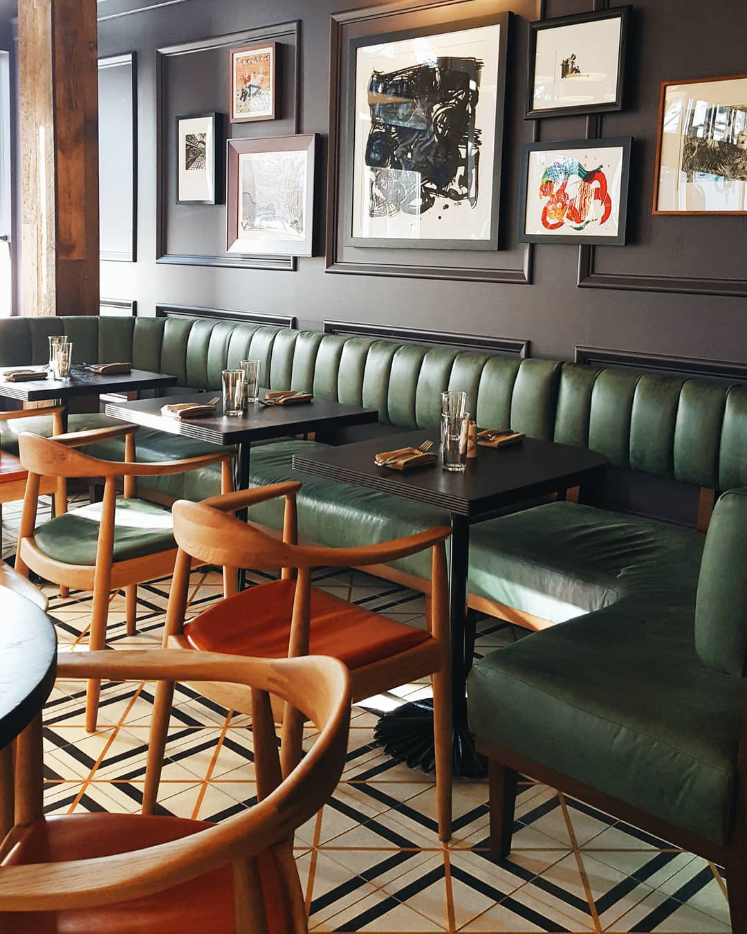 A stylish restaurant interior with green upholstered banquettes, wooden chairs with tan leather seats, black tables set formally, framed art on dark walls, and Brixton 8x8 tile flooring by vendor-unknown.