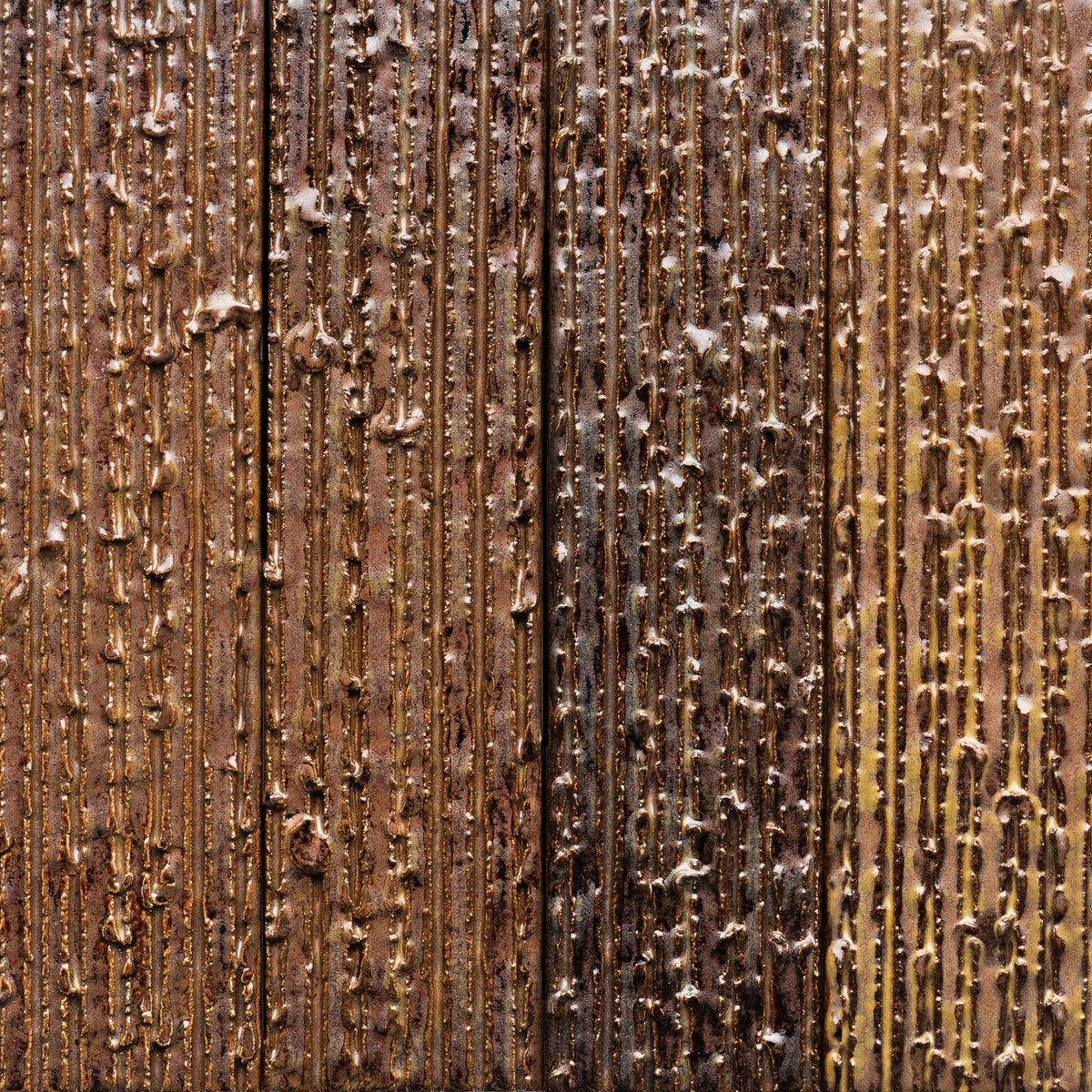 Close-up of Buckeye + Furrowed, Sample by Zia Tile shows a textured metal surface with vertical lines and raised bumps in brown, copper, and gold tones. Light reflects off the uneven surface, emphasizing its metallic texture.