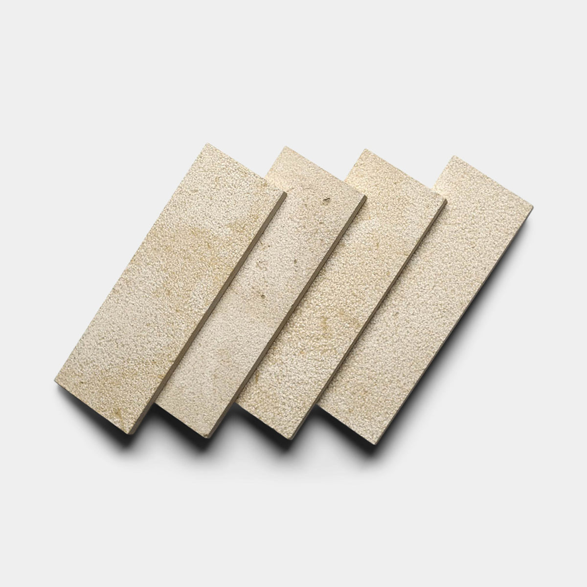 Four Buff 4x12 + Bush Hammered tiles by Zia Tile are arranged in a slightly overlapping diagonal row on a plain white background, casting soft shadows.
