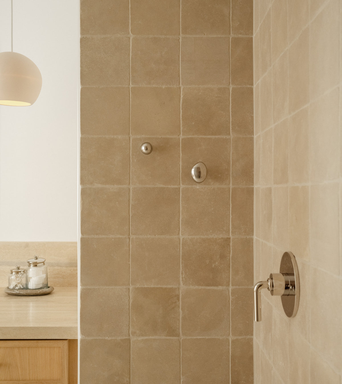 Minimalist bathroom featuring Zia Tile's Buff 6x6 + Honed tiles in beige, a modern round shower control, a countertop with glass jars, and a pendant light, all styled in neutral tones.