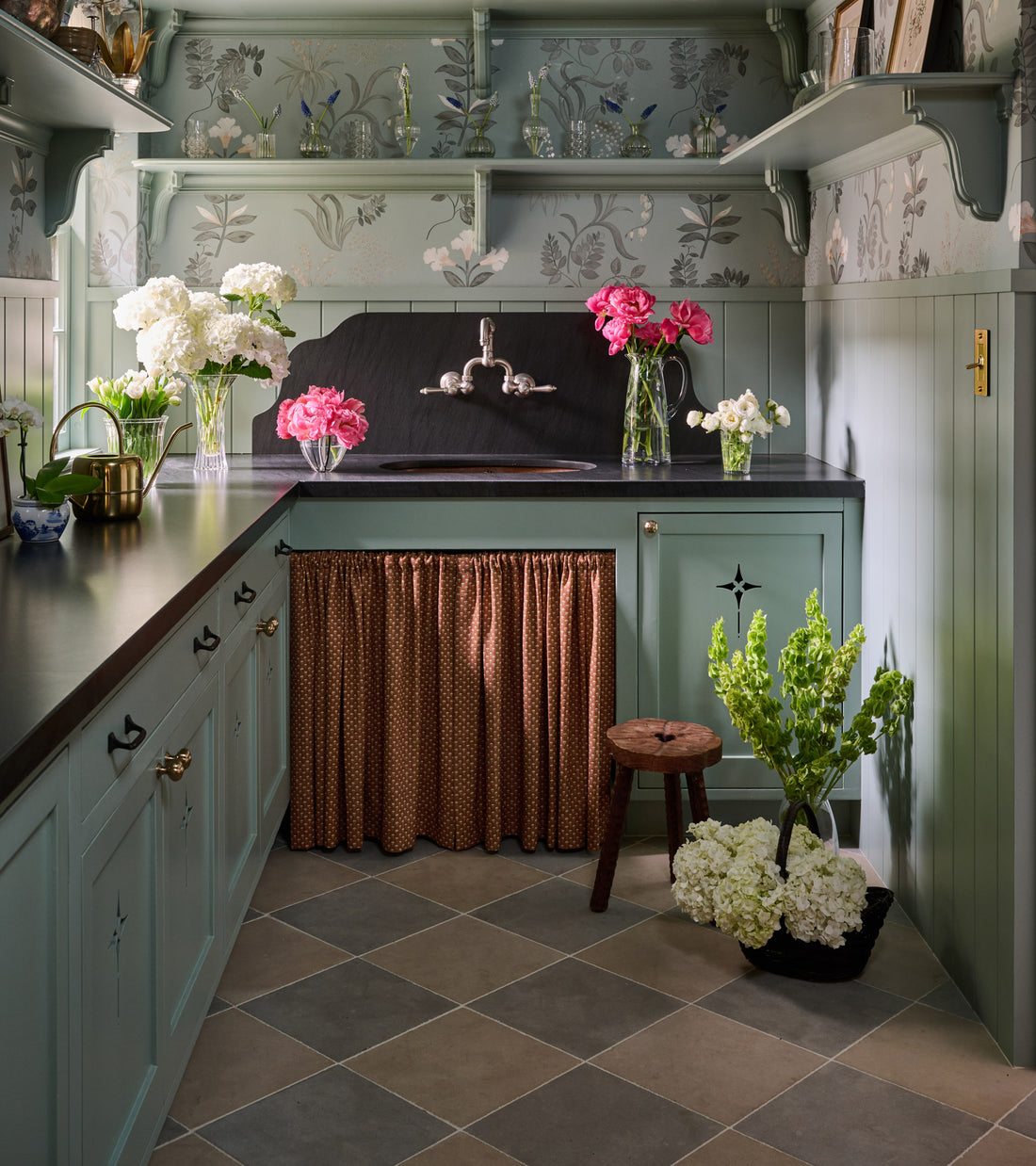 A vintage-style kitchen features sage green cabinets, black countertops, floral wallpaper, open shelves, and fresh flowers. Zia Tile’s Buff 12x12 + Honed stone tiles add texture beneath a brown skirted curtain under the sink.