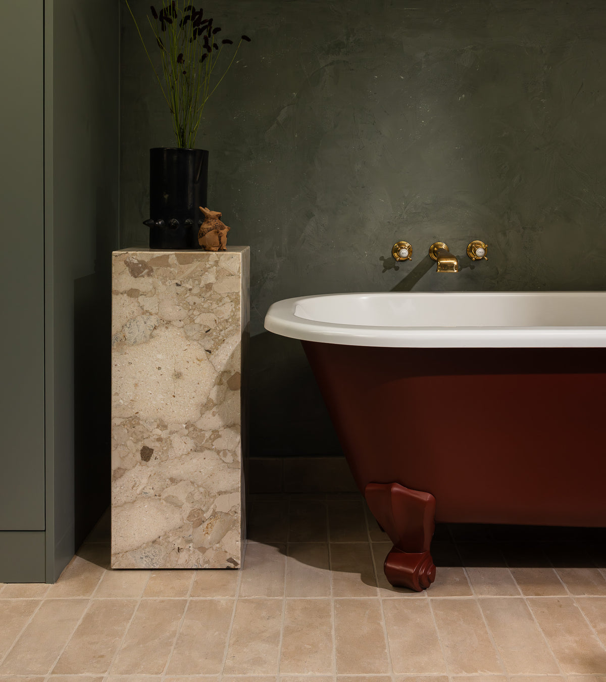 A stylish bathroom features a red clawfoot tub, terrazzo pedestal with black vase and decor, gold wall-mounted faucets, green textured walls, and beige Buff 4x12 + Honed tiles from Zia Tile adorning the floor.