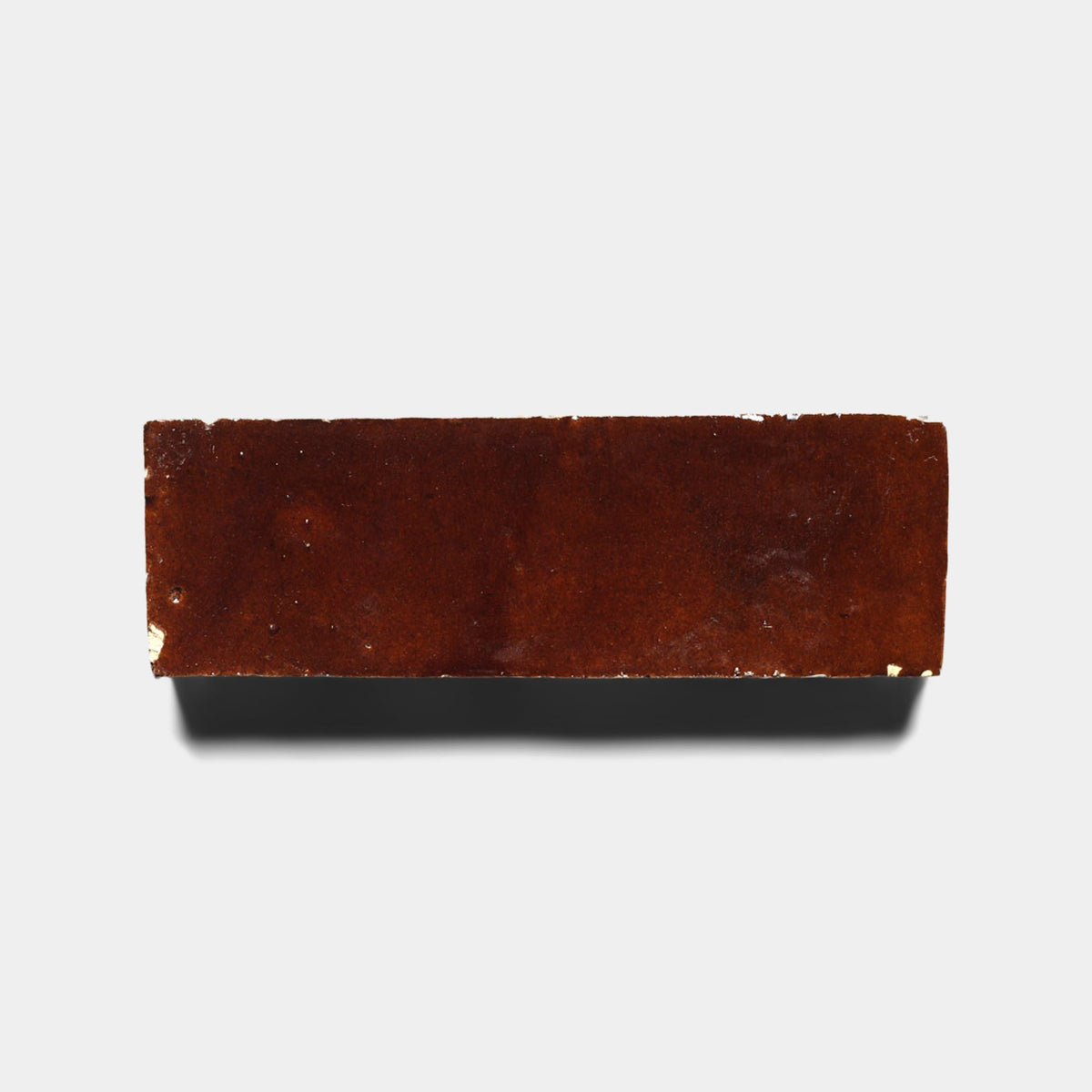A single rectangular Burnt Sugar 2x6 tile sample by Zia Tile, featuring a smooth brown handcrafted surface, casts a shadow on a plain white background.