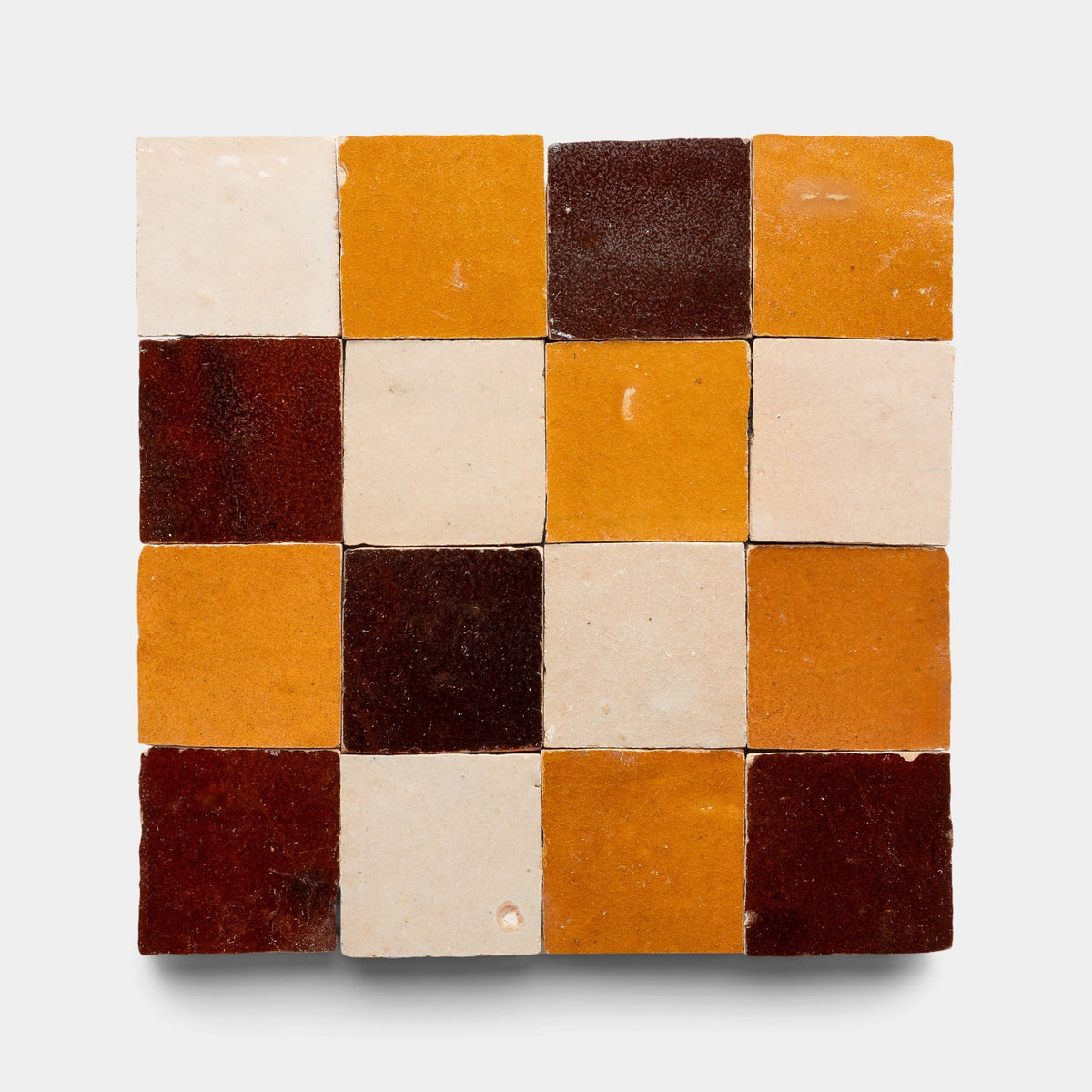 A sample of Burnt Sugar + Glazed Earth + Amber Checkerboard tiles by Zia Tile, featuring a 4x4 square grid of handmade cement tiles in brown, yellow-orange, and off-white on a plain light background.