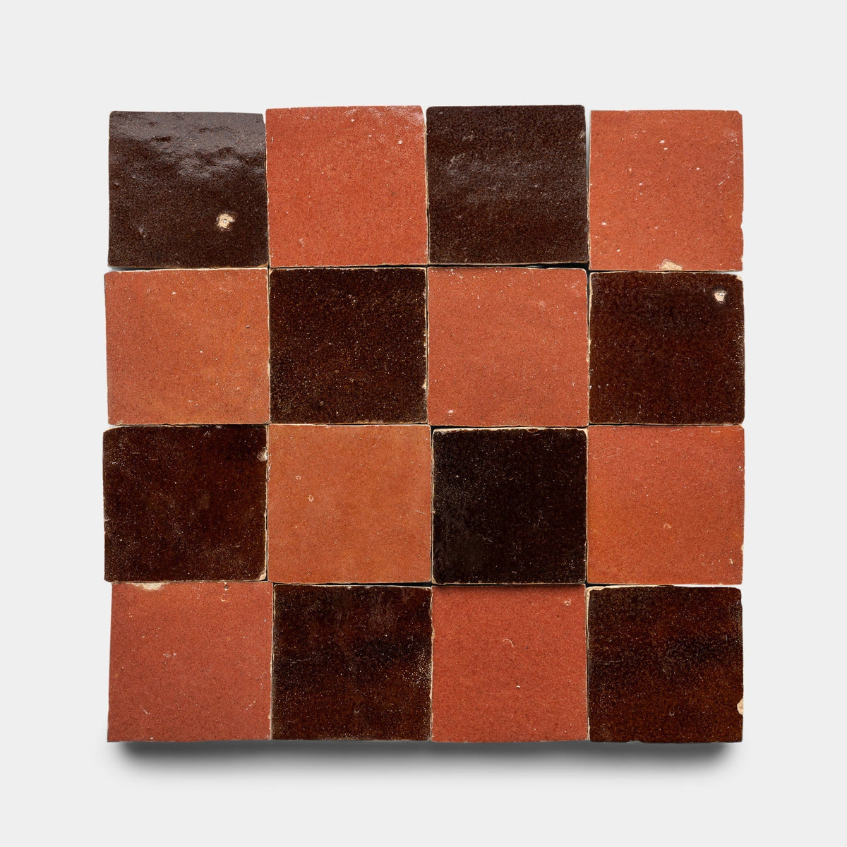 The Zia Tile Burnt Sugar + Terra Rosa Checkerboard Sample features 16 handmade tiles in a square layout, alternating dark brown and reddish-brown tones for a classic checkerboard effect against a light background.