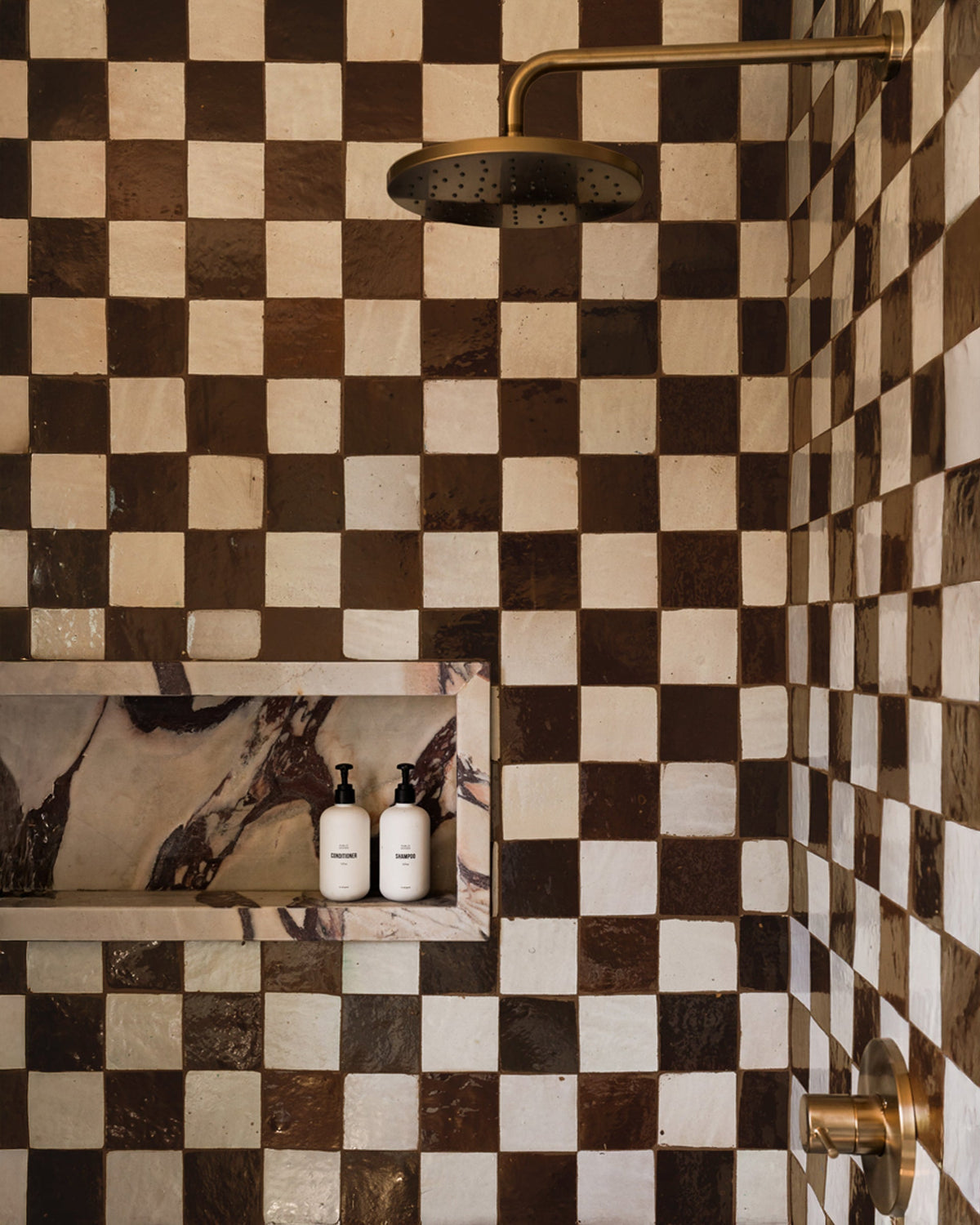 A shower featuring Zia Tile Burnt Sugar 2x6 tiles in brown and white, brass fixtures, and a built-in shelf holding two white bottles with black pumps. The shelf has a marbled background.