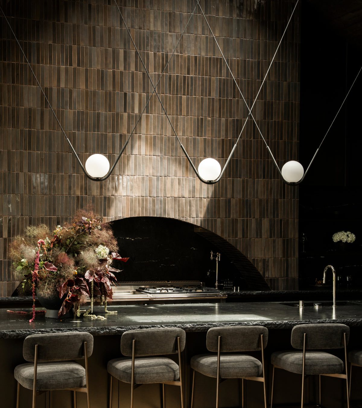 A dark kitchen featuring a long counter with bar stools, a Cacao Mini Bar tiled backsplash, and three globe pendant lights suspended above.