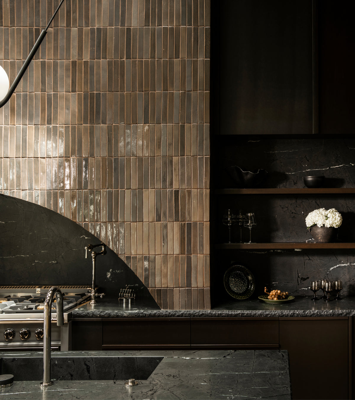 A kitchen wall tiled with vertical, brown-toned Cacao Mini Bar rectangular tiles next to dark stone surfaces and open shelving.