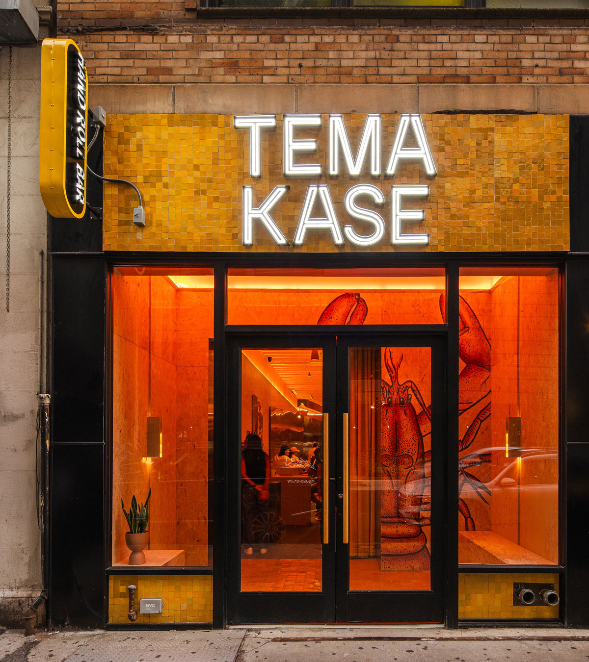 Street view of a restaurant featuring Zia Tile’s Cadmium 2x2 tiles, a neon TEMA KASE sign, and a large lobster mural near the entrance, all bathed in warm orange light.