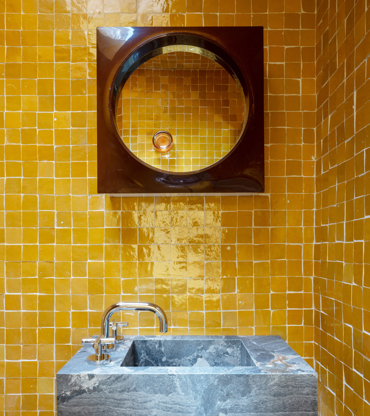 A modern bathroom features glossy orange Cadmium 2x2 tiles by Zia Tile, a circular mirror with a square black frame above a gray stone sink, and a shiny metal faucet.