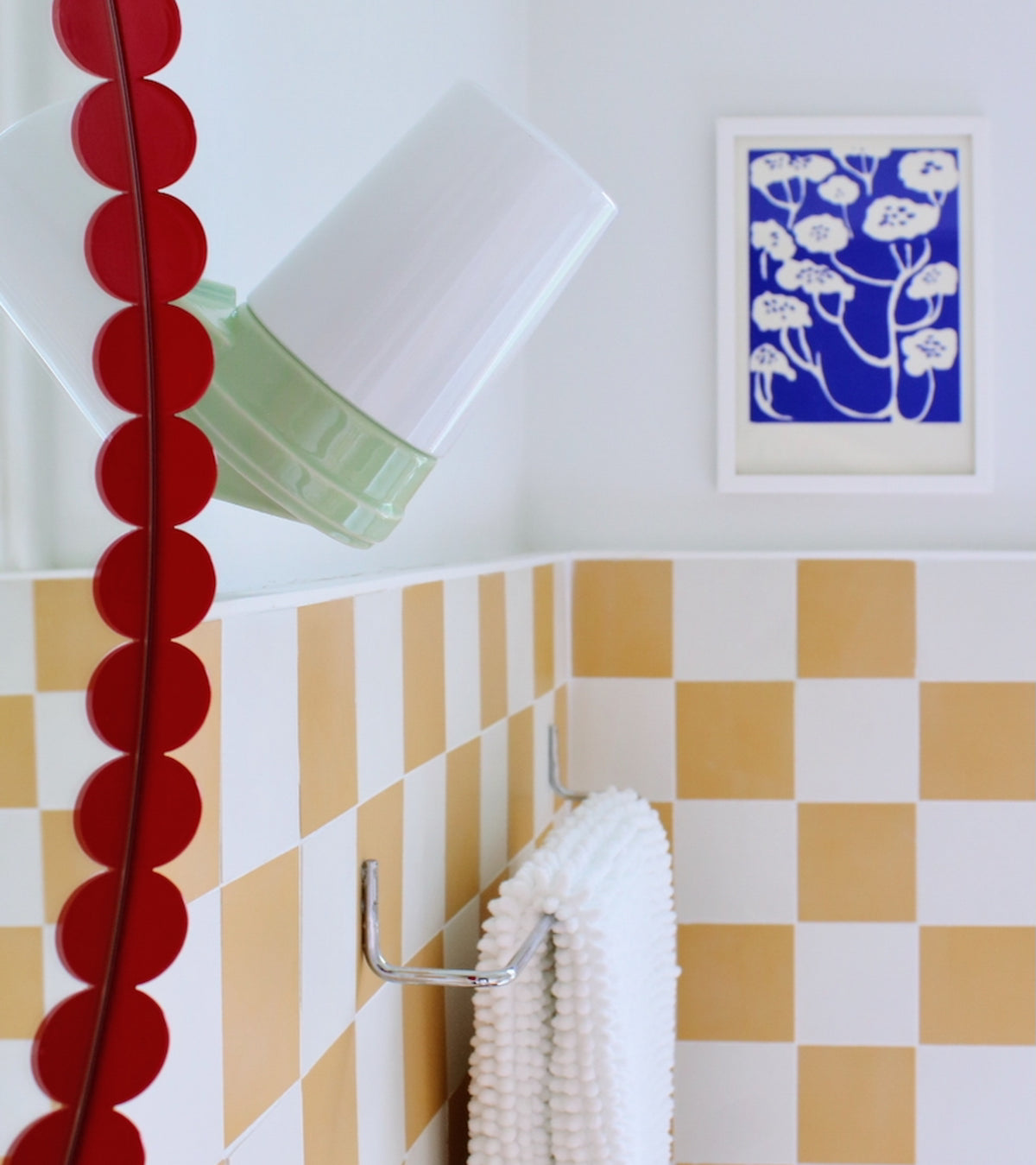 A bathroom features Cadmium 4x4 yellow and white checkered cement tile by vendor-unknown, a textured white towel on a silver rack, a red scalloped-edge mirror, modern white lamp, and blue botanical print—perfect kitchen backsplash inspiration.