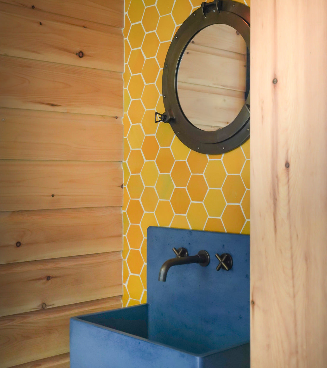 A bathroom showcases a round mirror, a yellow Cadmium Hex tile wall by Zia Tile, a modern black faucet, and a blue rectangular sink, all framed by light wooden paneling.