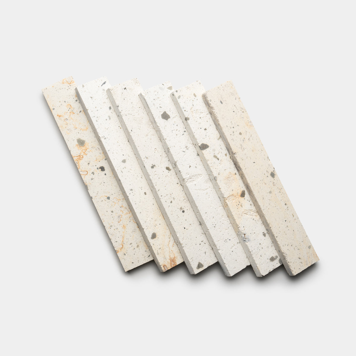 Six Zia Tile Caldera 2x12 rectangular terrazzo tiles, featuring a white base with scattered grey and beige speckles, are arranged in a slightly fanned-out pattern on a plain white background.