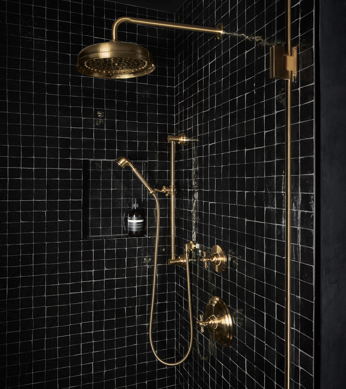 A sleek, luxurious shower showcases Zia Tile’s Carbon Black 2x2 tiles, accented by gold fixtures—a rain showerhead, handheld shower, faucet, and controls. A built-in shelf displays a single dark bottle.
