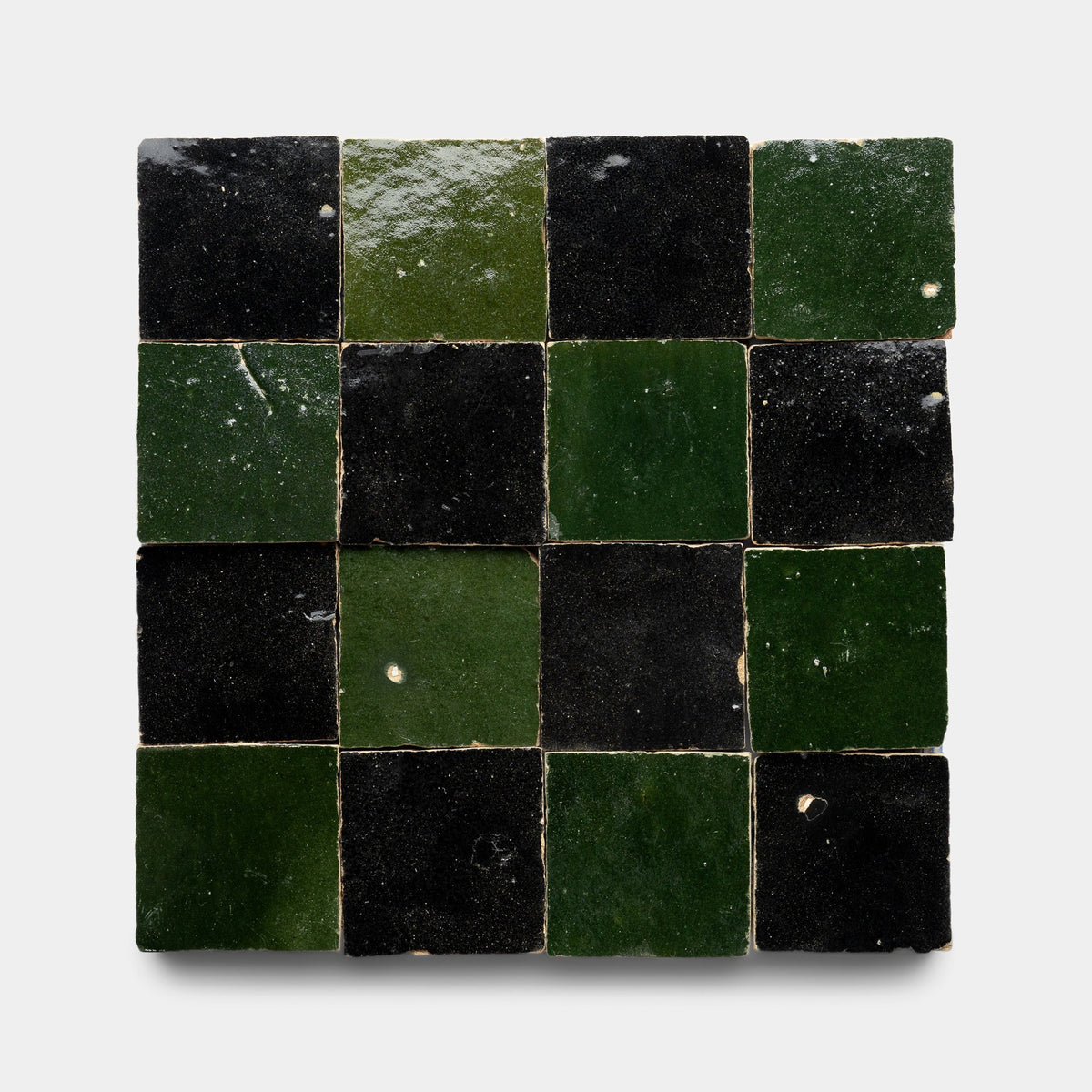 The Carbon Black + Racing Green Checkerboard sample by Zia Tile features sixteen glossy handmade tiles arranged in a square, alternating dark green and black for a checkerboard effect on a light background.