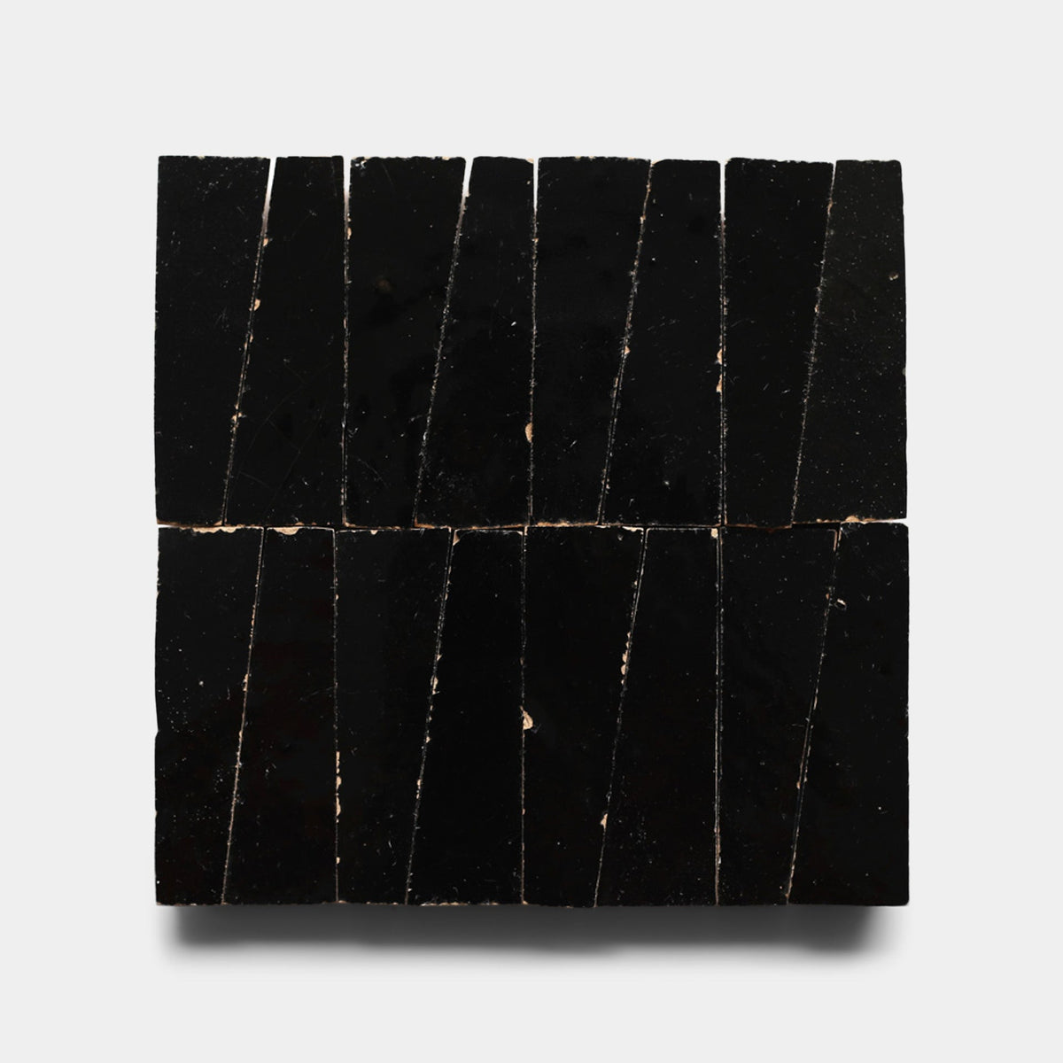 Eight Carbon Black Trapezoid samples by Zia Tile, handmade with uneven edges and diagonal cuts, are arranged vertically in a square on a light gray background. The glossy black tiles feature small edge chips.