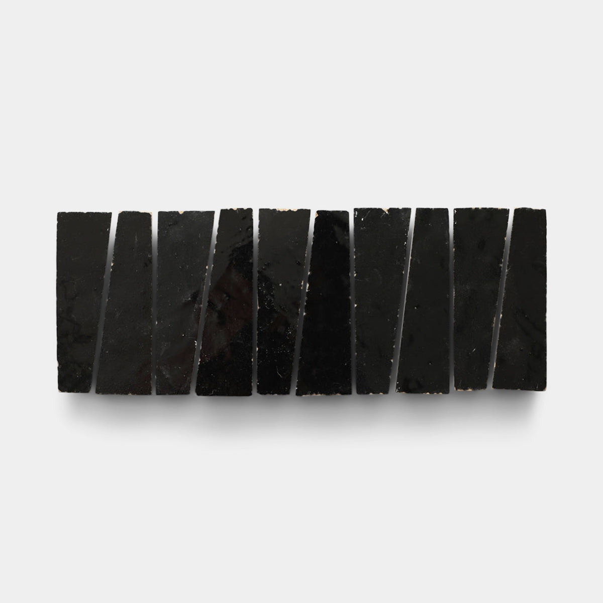 Ten Carbon Black Trapezoid sample tiles by Zia Tile, glossy and uniquely shaped, are arranged horizontally on a white background. Each handcrafted tile slightly differs in width and angle for an artistic, irregular effect.