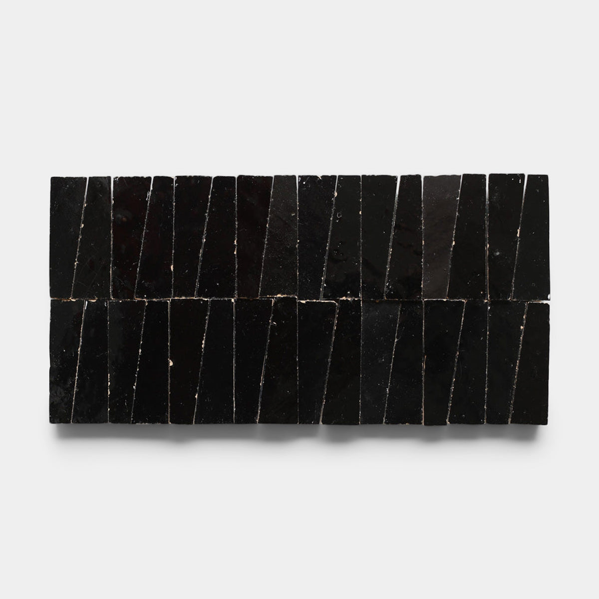 Two rows of glossy, black Carbon Black Trapezoid handmade tiles by Zia Tile, each with irregular shapes and uneven edges, are arranged vertically and closely together on a plain white background.
