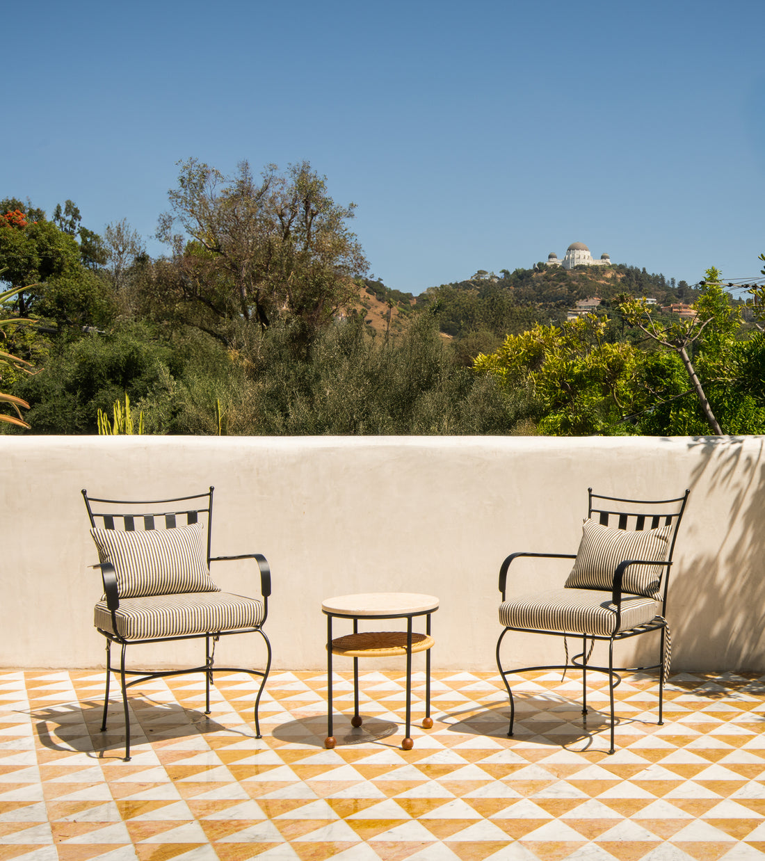 Two metal chairs with striped cushions and a small round table rest on a Zia Tile Isola in Carrara + Giallo Reale patio, with trees behind and a domed observatory on a distant hill beneath a clear blue sky.