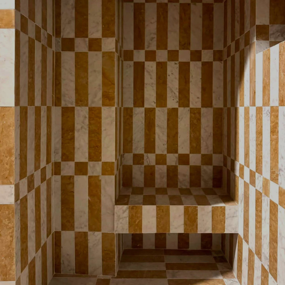 A room features walls, floor, and a bench clad in Zia Tile’s Lucca in Carrara + Giallo Reale cement tiles, forming a striking checkerboard design for an optical illusion effect.