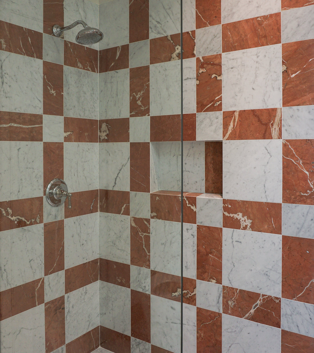 A shower interior features Zia Tile’s Etna in Carrara + Rosso Alicante handmade tiles in a red and white checkerboard pattern, with a silver showerhead, handle, and built-in storage niche.