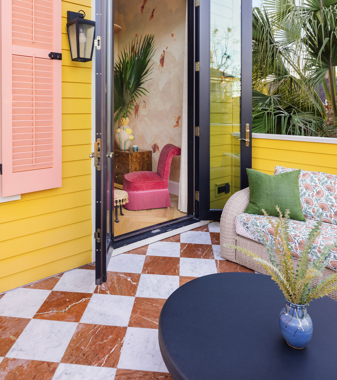 A vibrant yellow wall with pink shutters opens to a cozy room. The patio features Zia Tile’s Piazza in Carrara + Rosso Alicante checkered cement tiles, a wicker sofa with green pillows, and a blue vase on a black table.