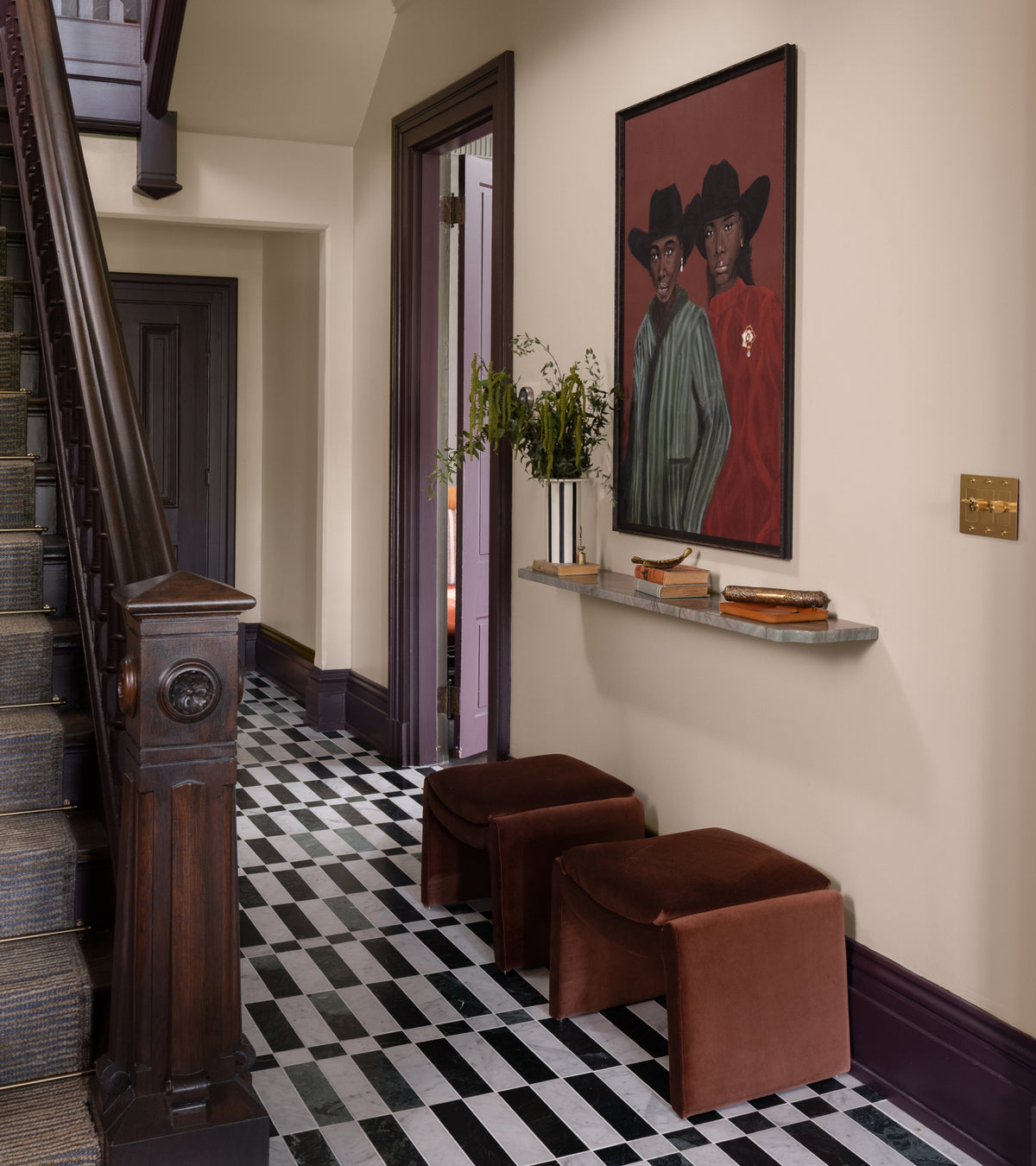 A hallway with a geometric, handmade Lucca in Carrara + Verde Alpi tile floor, two brown stools, and a painting of two people in hats hanging above a narrow wall-mounted shelf.