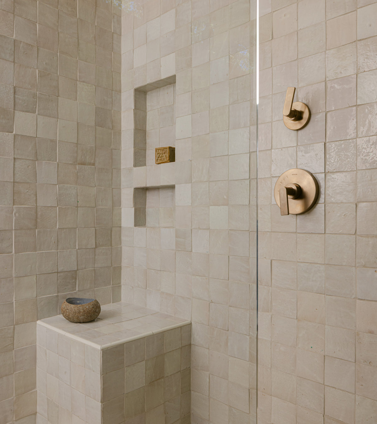 A shower features Casablanca 4x4 beige tiles by vendor-unknown, two built-in shelves with a small brown object, a tiled bench holding a small round bowl, and modern brass controls on the wall.