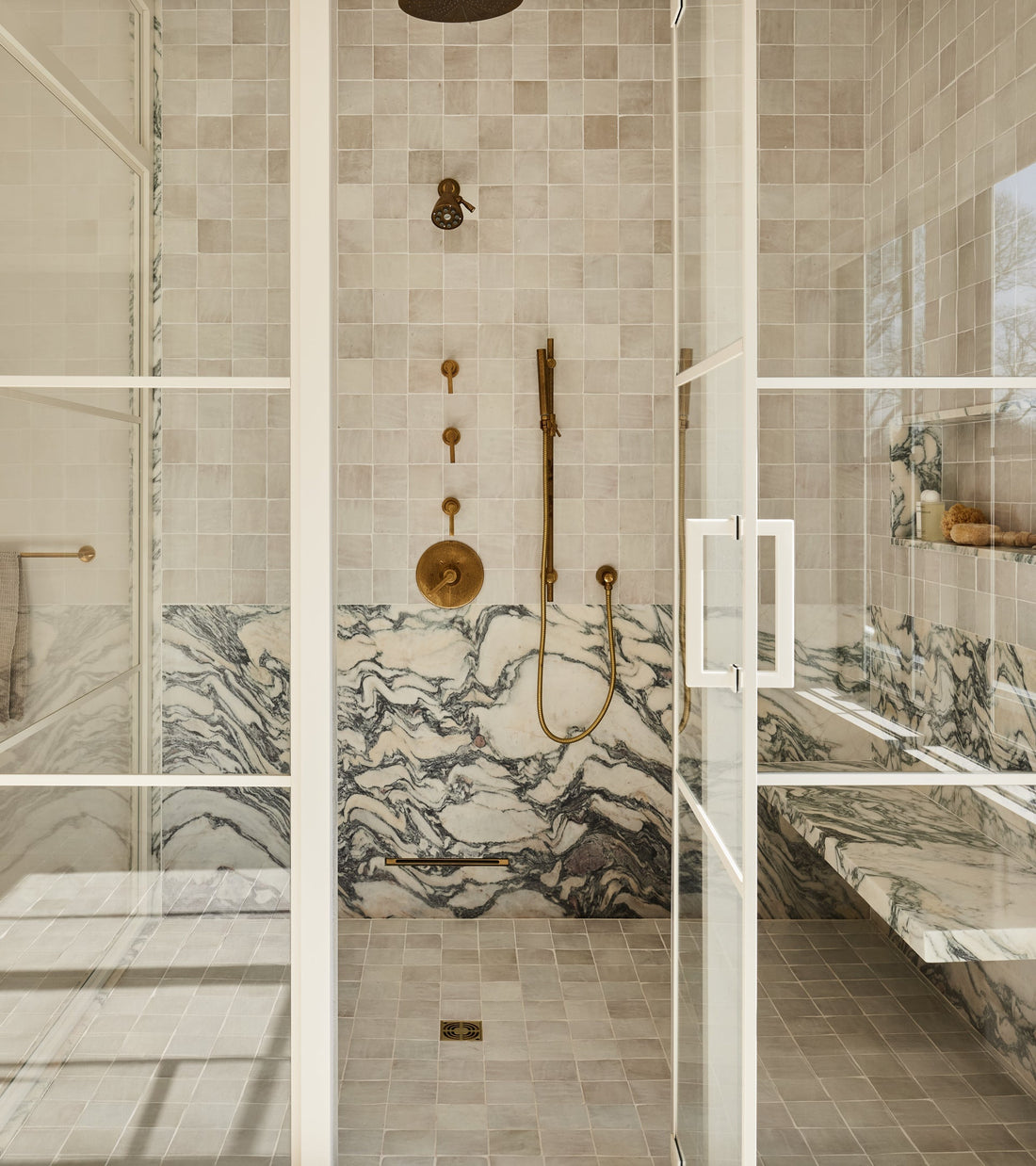 Modern walk-in shower features Casablanca 4x4 beige tiles by vendor-unknown, glass doors, brass fixtures, and gray-veined marble walls. A built-in shelf holds toiletries as natural light pours in through a window.