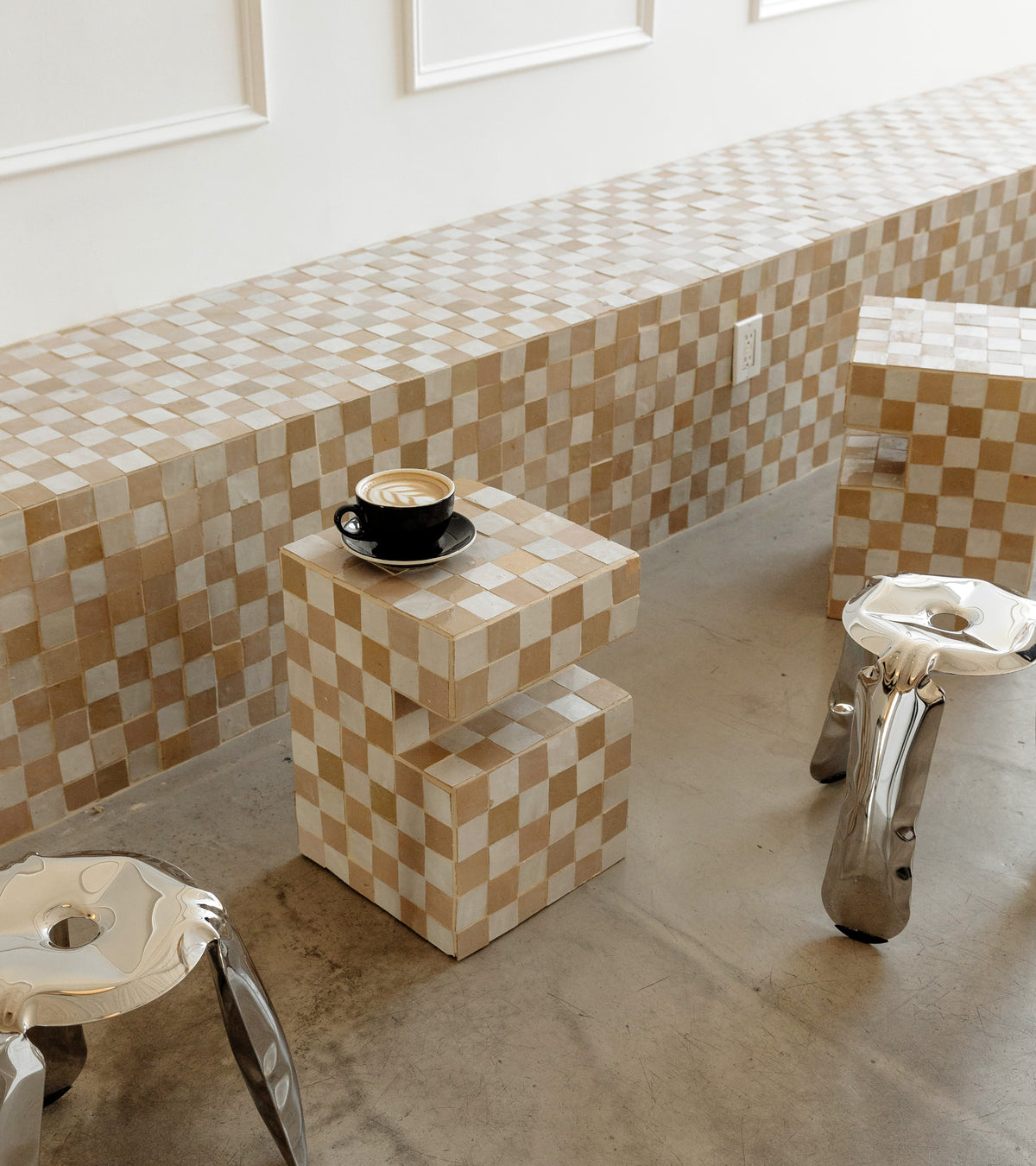 A cappuccino in a black cup sits on a small Casablanca 2x2 zellige cube, near a matching long tiled bench and two silver stools.