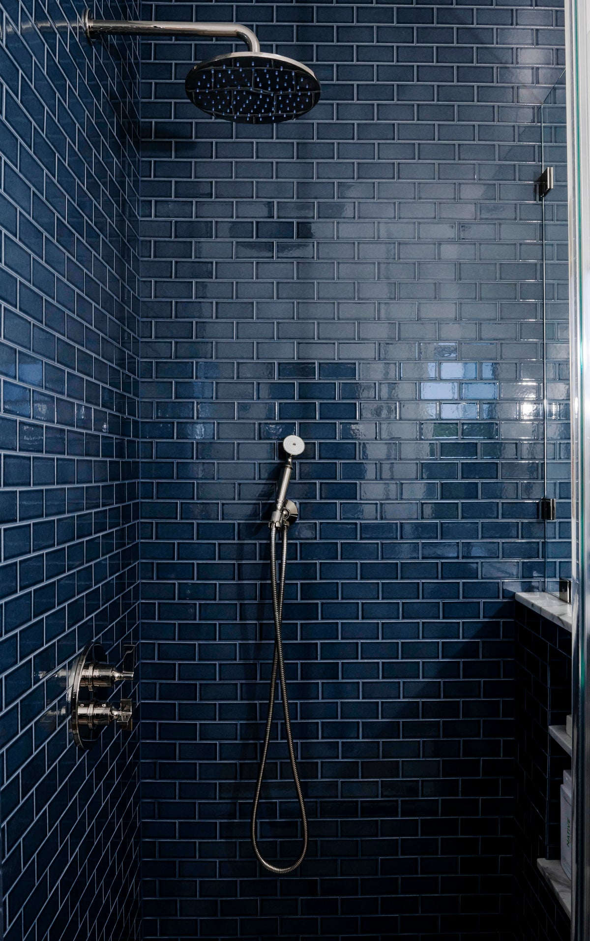 A modern shower featuring Zia Tile's Ethereal Blue 2x4 ceramic subway tiles, a ceiling-mounted round rainfall showerhead, a handheld showerhead with hose, and built-in architectural-grade metal fixtures.