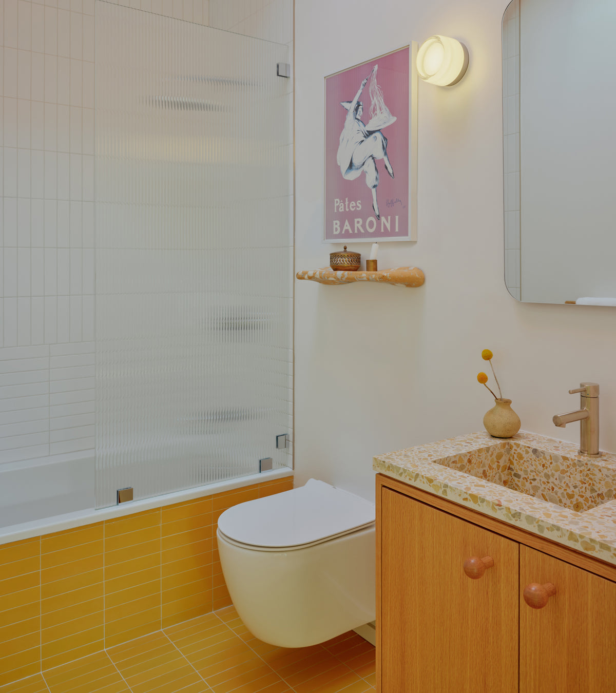 A bathroom featuring Citron 2x8 matte ceramic tiles in mustard yellow on the floor and bathtub surround, a floating wooden vanity with a terrazzo-style countertop, and a pink Pâtes Baroni poster on the wall.