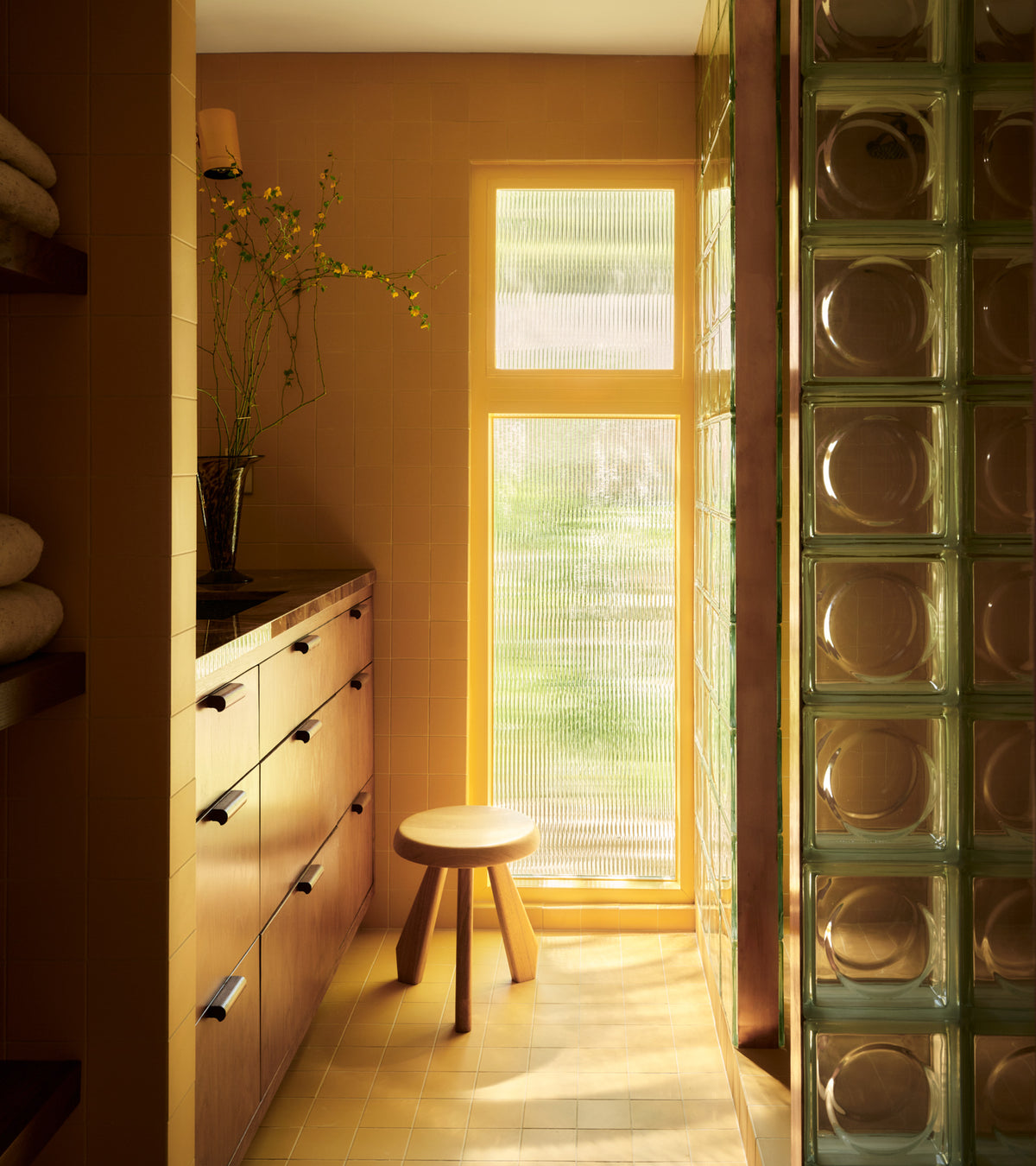 A sunlit bathroom features Zia Tile's Citron 4x4 ceramics in cheerful yellow, a wooden cabinet and round stool, folded towels on shelves, and a glass block partition by a textured window with green-tinted outdoor light streaming through.