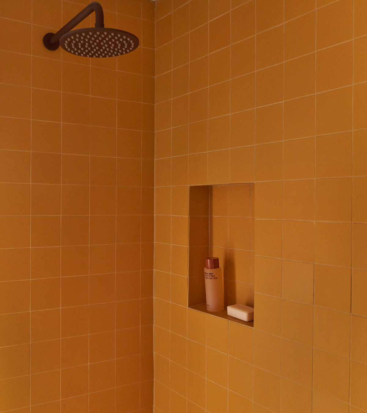 A shower features Zia Tile's Citron 4x4 ceramic tiles, a round rainfall showerhead, and a recessed shelf with a bottle and bar of soap.