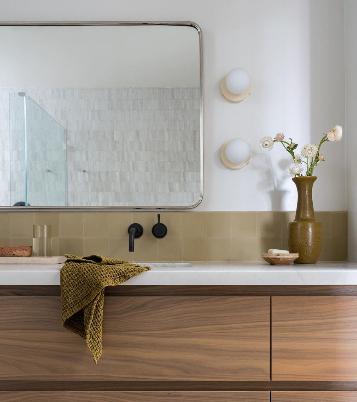 A modern bathroom features a wooden vanity, beige tiles, a rectangular mirror flanked by round wall lights, black faucet, green towel, and brown vase with white flowers atop the counter. The backsplash is Clay 4x4 tile by vendor-unknown.