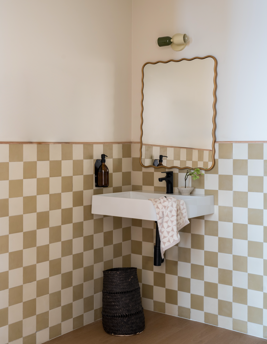 A small corner sink with a black faucet, patterned towel, brown bottle, and green plant sits below a wavy mirror. Walls showcase vendor-unknown’s Clay 4x4 beige and white tile; a wicker basket rests on the floor.