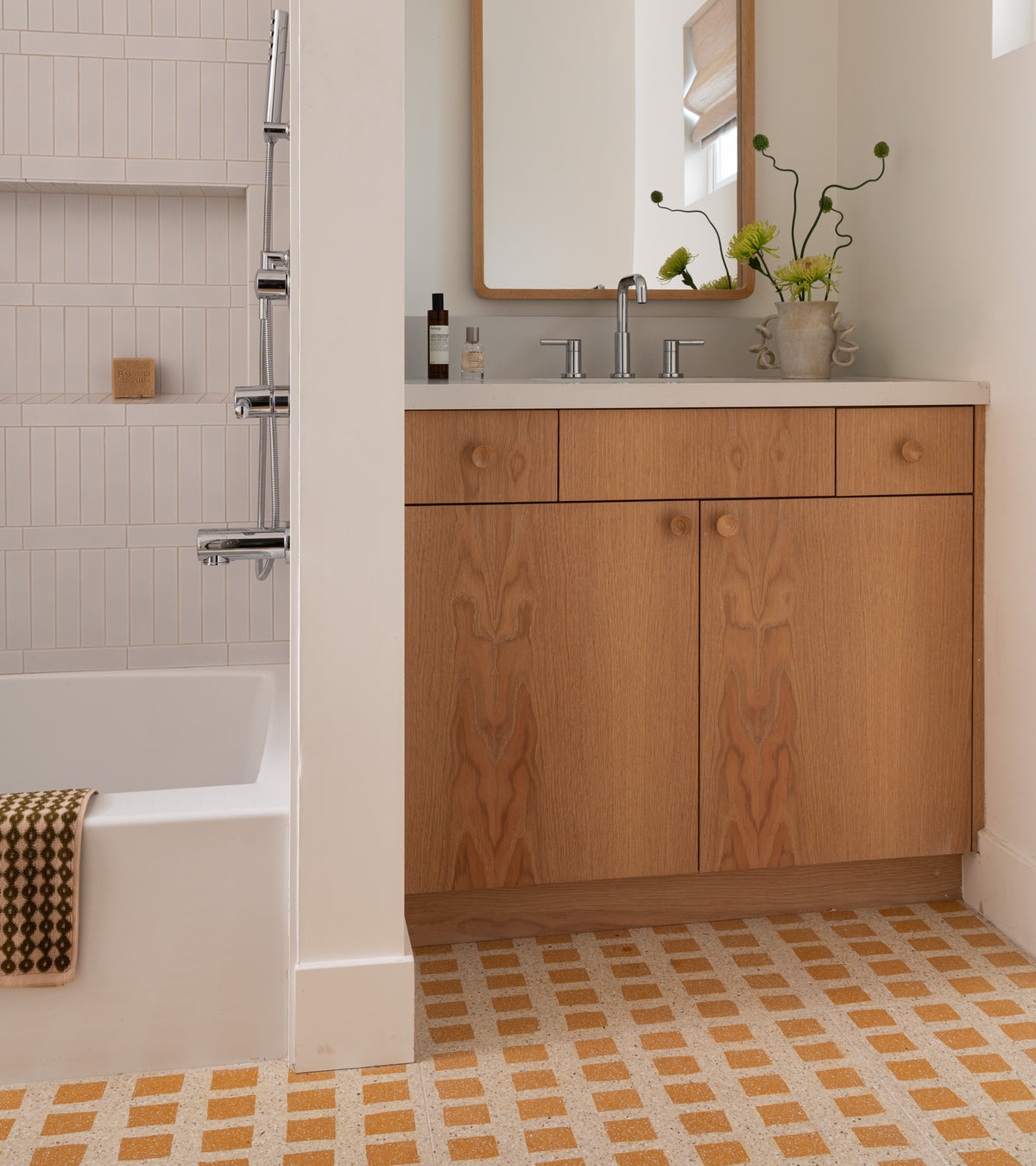 A modern bathroom with a white bathtub, wall-mounted shower, wooden vanity, rectangular mirror, green flower vase, and Zia Tile's Constellation Bone + Ginger 24x24 terrazzo cement tile flooring in a yellow and white checkered pattern.