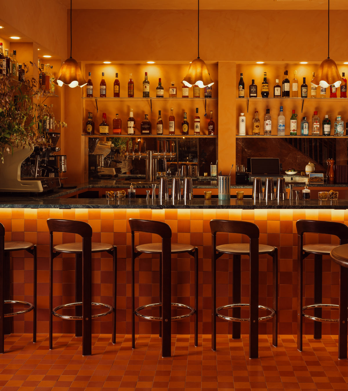 A warm bar interior highlights a Coral 4x4 tile counter by vendor-unknown, paired with high stools, pendant lights, and shelves of bottles and glassware against an orange-toned wall.