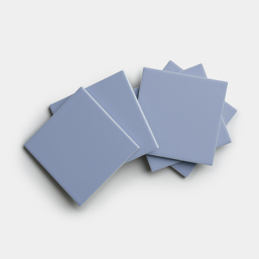 Five Cornflower 4x4 square ceramic tiles from Zia Tile overlap on a white background, showcasing their light blue color.
