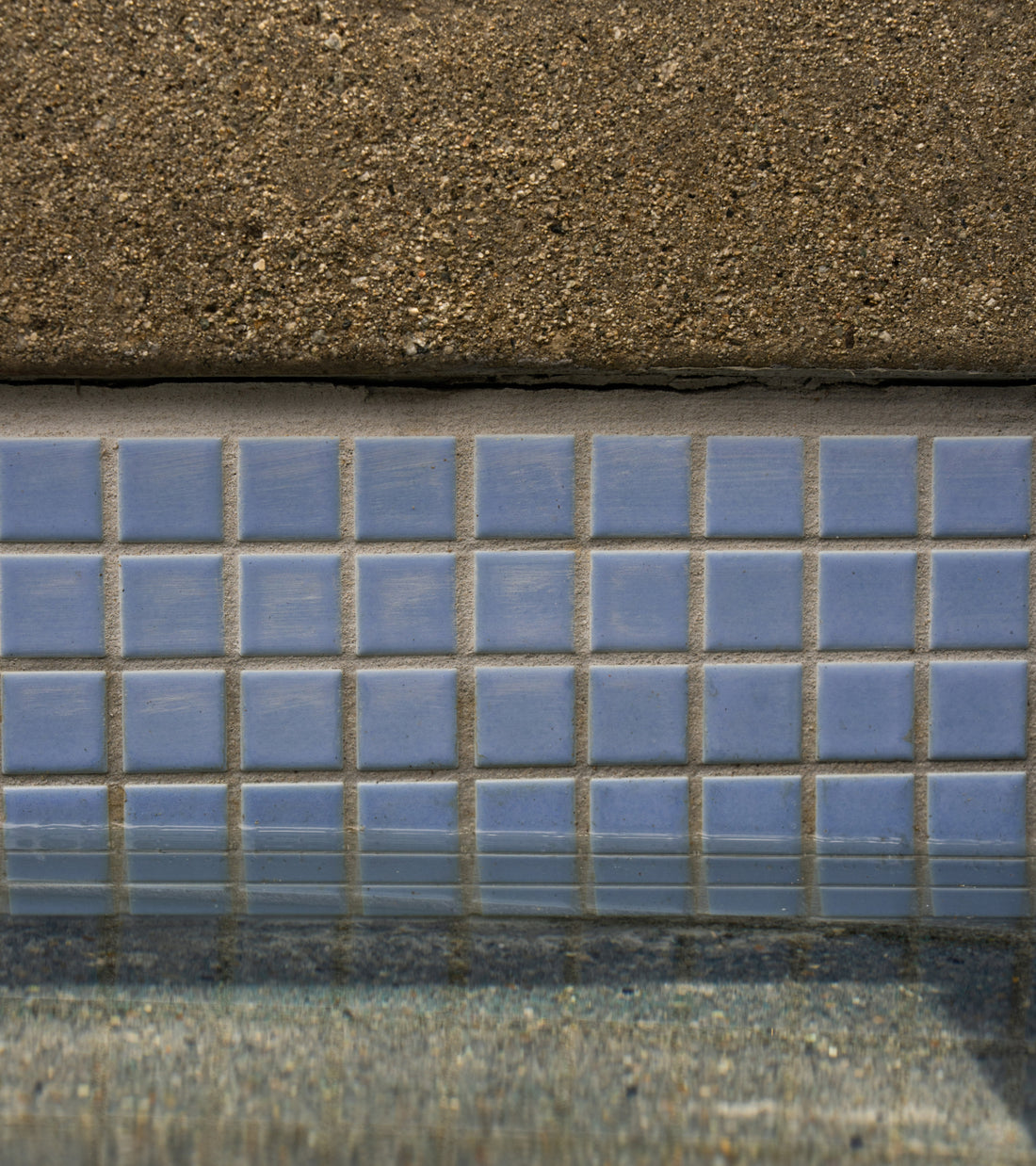 A row of Cornflower 1x1 light blue square tiles bordering the edge of a swimming pool below a concrete deck.